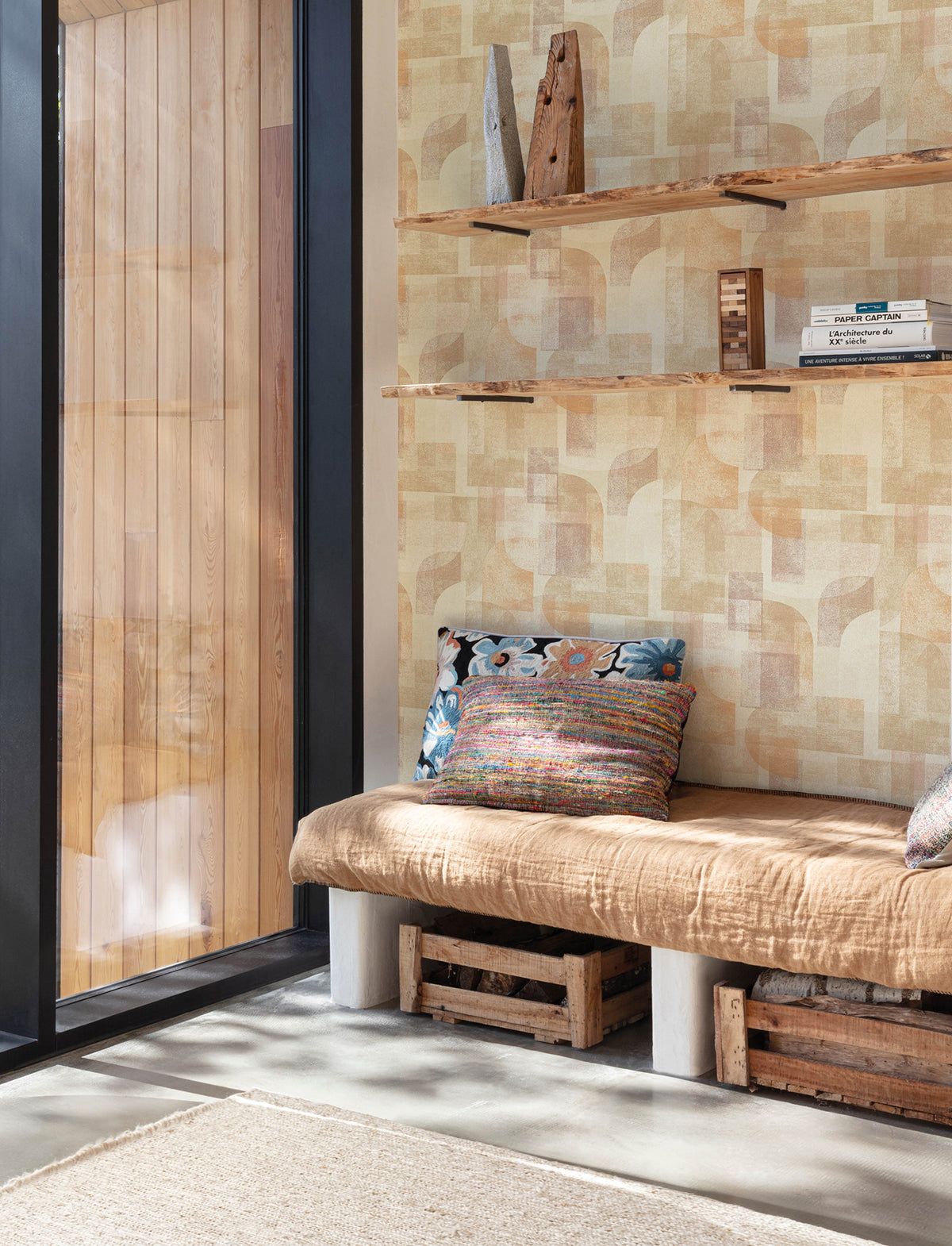 A cozy nook with a tan cushioned bench, colorful pillows, wooden crate storage underneath, open shelves filled with books and rustic decor, next to a large window framed by natural light and accented with Chesapeake Cassandra Block Geometric Wallpaper by York Wallcoverings.
