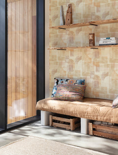 A cozy nook with a tan cushioned bench, colorful pillows, wooden crate storage underneath, open shelves filled with books and rustic decor, next to a large window framed by natural light and accented with Chesapeake Cassandra Block Geometric Wallpaper by York Wallcoverings.