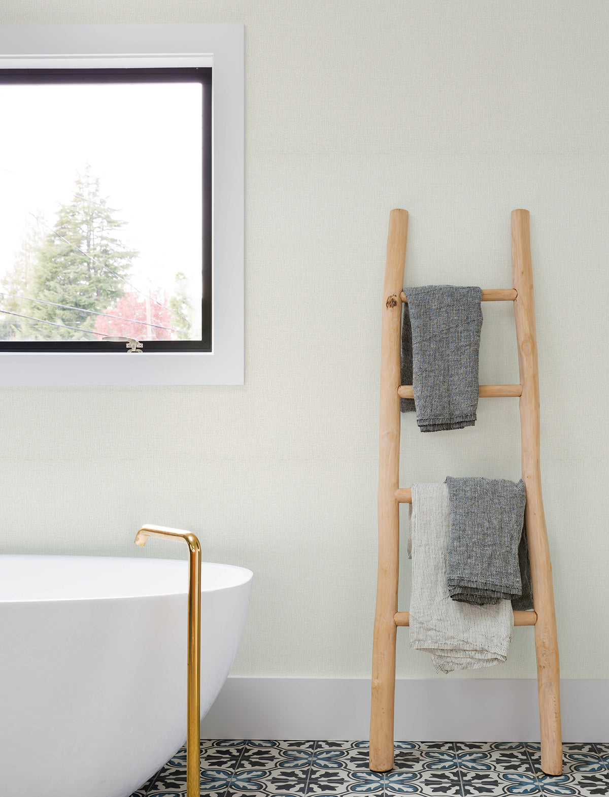 A minimalist bathroom featuring York Wallcoverings’ Chesapeake Sonia Faux Linen Wallpaper, a white tub with a gold faucet, a patterned tile floor, a wooden ladder displaying gray and white towels, and a window showcasing tree views.