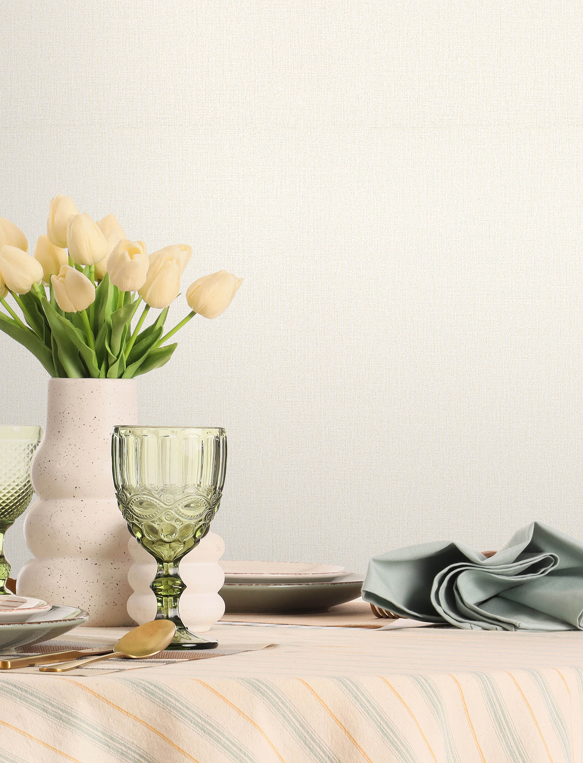 A table set with plates, a green glass, a folded napkin, and a vase of pale yellow tulips on a striped tablecloth is placed against a plain light background featuring Chesapeake Sonia Faux Linen Wallpaper by York Wallcoverings.