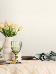 A table set with plates, a green glass, a folded napkin, and a vase of pale yellow tulips on a striped tablecloth is placed against a plain light background featuring Chesapeake Sonia Faux Linen Wallpaper by York Wallcoverings.
