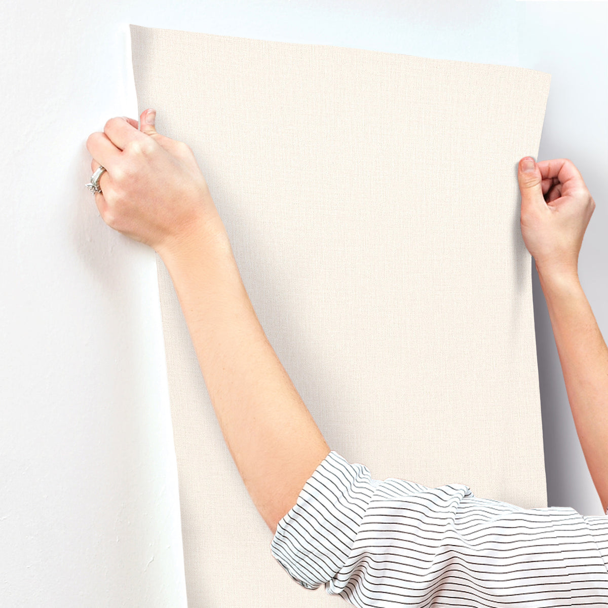 A person in a striped shirt applies Chesapeake Sonia Faux Linen Wallpaper by York Wallcoverings to a white wall, smoothing it with both hands.