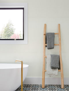 A wooden ladder holding three folded towels stands next to a white bathtub with a gold faucet, near a window and decorative tile floor in a bright bathroom accented with Chesapeake Sonia Faux Linen Wallpaper by York Wallcoverings.
