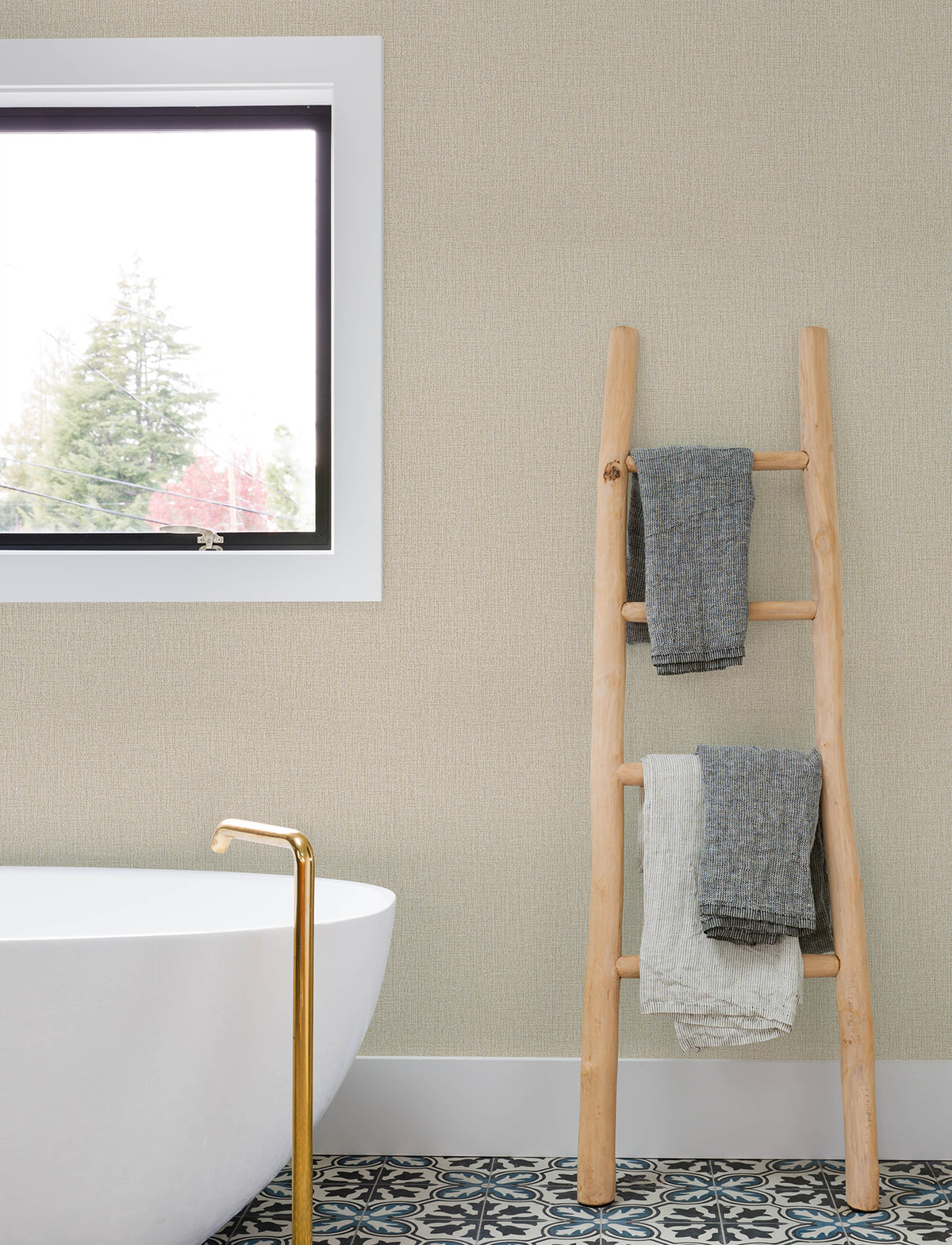 A light wood ladder with gray and white towels leans against a wall covered in Chesapeake Sonia Faux Linen Wallpaper by York Wallcoverings, next to a white bathtub with a gold faucet. A window reveals trees outside, and patterned tile covers the bathroom floor.