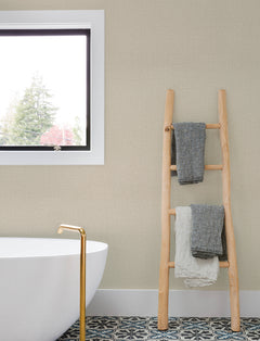 A light wood ladder with gray and white towels leans against a wall covered in Chesapeake Sonia Faux Linen Wallpaper by York Wallcoverings, next to a white bathtub with a gold faucet. A window reveals trees outside, and patterned tile covers the bathroom floor.