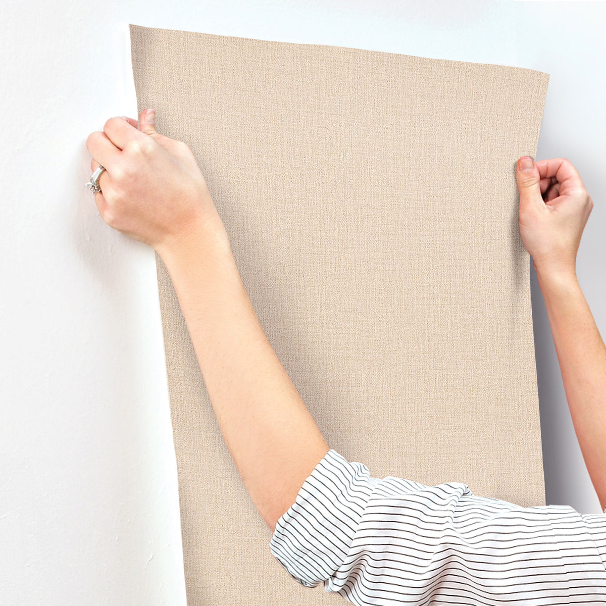 A person in a striped shirt is applying Chesapeake Sonia Faux Linen Wallpaper by York Wallcoverings, featuring a beige textured finish, to a white wall and smoothing it with their hands.