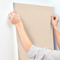 A person in a striped shirt is applying Chesapeake Sonia Faux Linen Wallpaper by York Wallcoverings, featuring a beige textured finish, to a white wall and smoothing it with their hands.