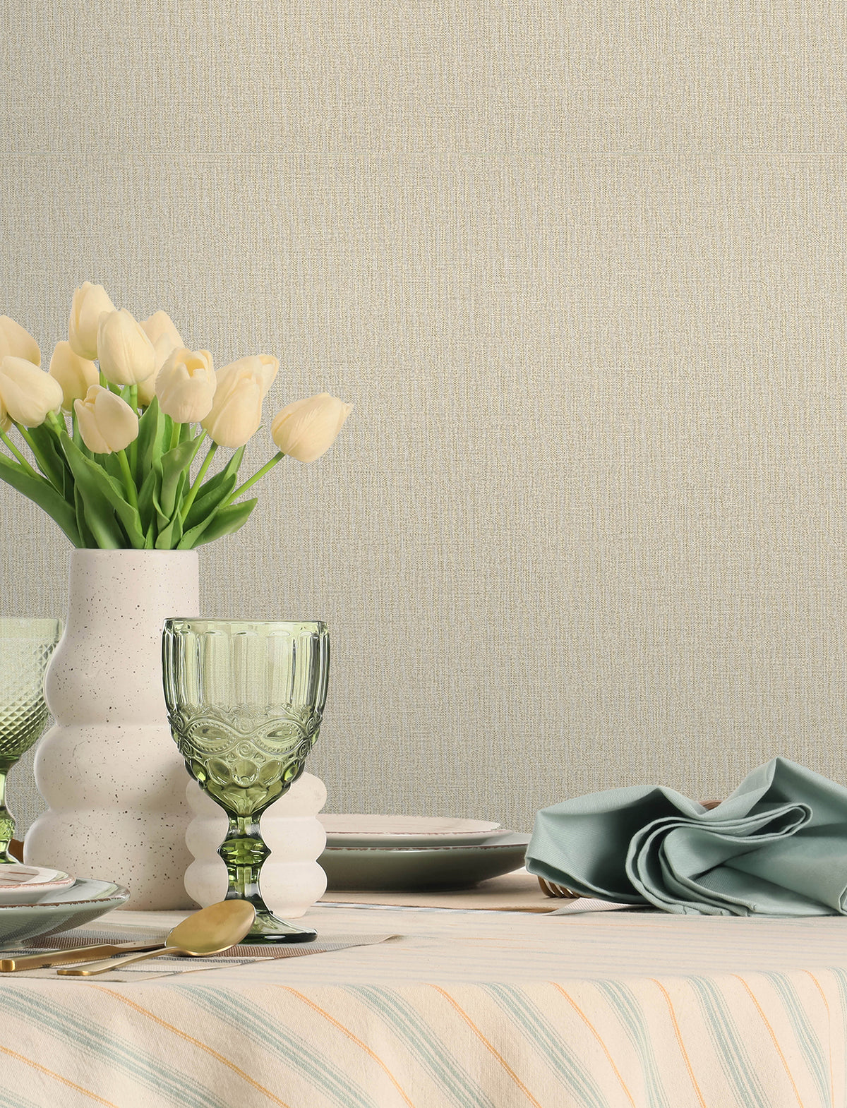 A table set with green glassware, a white vase of pale yellow tulips, plates, and a folded blue napkin on a striped tablecloth sits against a wall adorned with Chesapeake Sonia Faux Linen Wallpaper by York Wallcoverings.