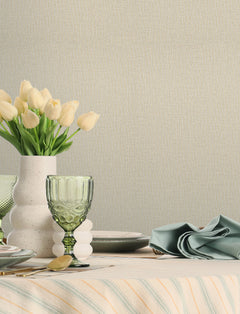 A table set with green glassware, a white vase of pale yellow tulips, plates, and a folded blue napkin on a striped tablecloth sits against a wall adorned with Chesapeake Sonia Faux Linen Wallpaper by York Wallcoverings.