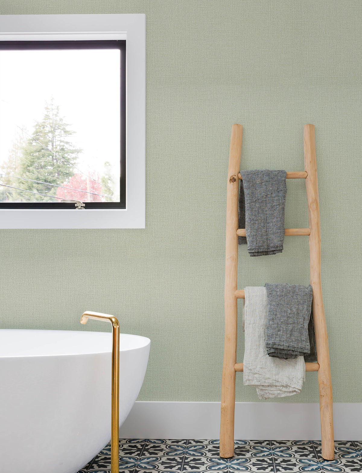 A modern bathroom featuring a white tub, gold faucet, patterned floor tiles, and a wooden ladder holding folded towels sits against walls adorned with Chesapeake Sonia Faux Linen Wallpaper by York Wallcoverings, beside a window that reveals trees outside.