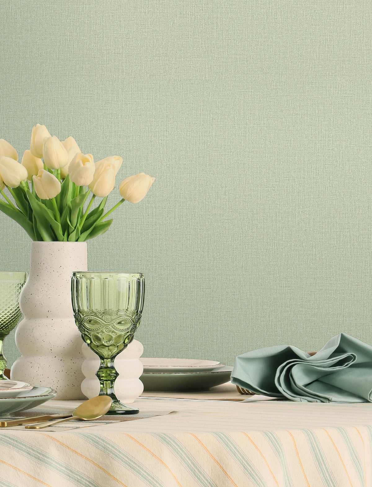 A table set with green glassware, a white vase of cream tulips, plates, cutlery, and a folded green napkin sits atop a striped tablecloth, set against walls adorned with Chesapeake Sonia Faux Linen Wallpaper by York Wallcoverings.