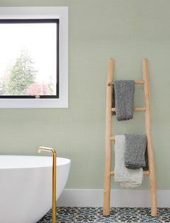 A modern bathroom with a white tub, gold faucet, patterned tile floor, a window, and a wooden ladder holding two towels sits against walls adorned with Chesapeake Sonia Faux Linen Wallpaper by York Wallcoverings.