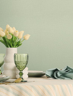 A table set with green glassware, a white vase of pale yellow tulips, plates, cutlery, and a folded green napkin is arranged against a pale green textured wall covered in York Wallcoverings’ Chesapeake Sonia Faux Linen Wallpaper.