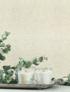 Two lit white candles in glass holders sit on a silver tray, surrounded by green eucalyptus branches, against a soft beige Chesapeake Tansy Distressed Wallpaper by York Wallcoverings.