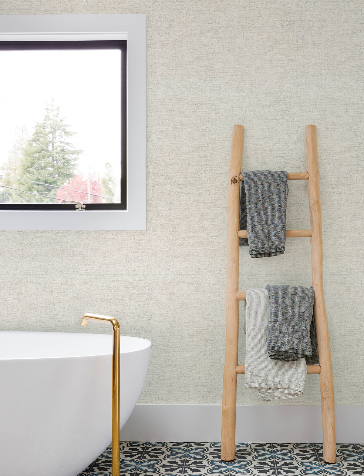 A modern bathroom featuring Chesapeake Tansy Distressed Wallpaper by York Wallcoverings, a white freestanding tub with a gold faucet, patterned tile flooring, a wooden ladder holding two towels, and a window framing views of trees outside.