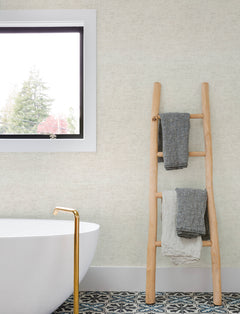 A modern bathroom featuring Chesapeake Tansy Distressed Wallpaper by York Wallcoverings, a white freestanding tub with a gold faucet, patterned tile flooring, a wooden ladder holding two towels, and a window framing views of trees outside.