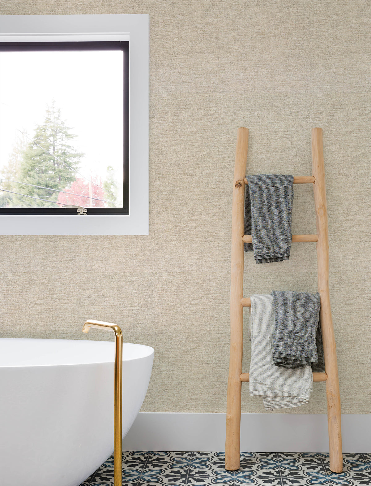 A wooden ladder holding three towels leans against a wall covered in Chesapeake Tansy Distressed Wallpaper by York Wallcoverings, next to a white bathtub with a gold faucet. A window and patterned tile floor are also visible.