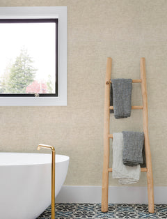 A wooden ladder holding three towels leans against a wall covered in Chesapeake Tansy Distressed Wallpaper by York Wallcoverings, next to a white bathtub with a gold faucet. A window and patterned tile floor are also visible.