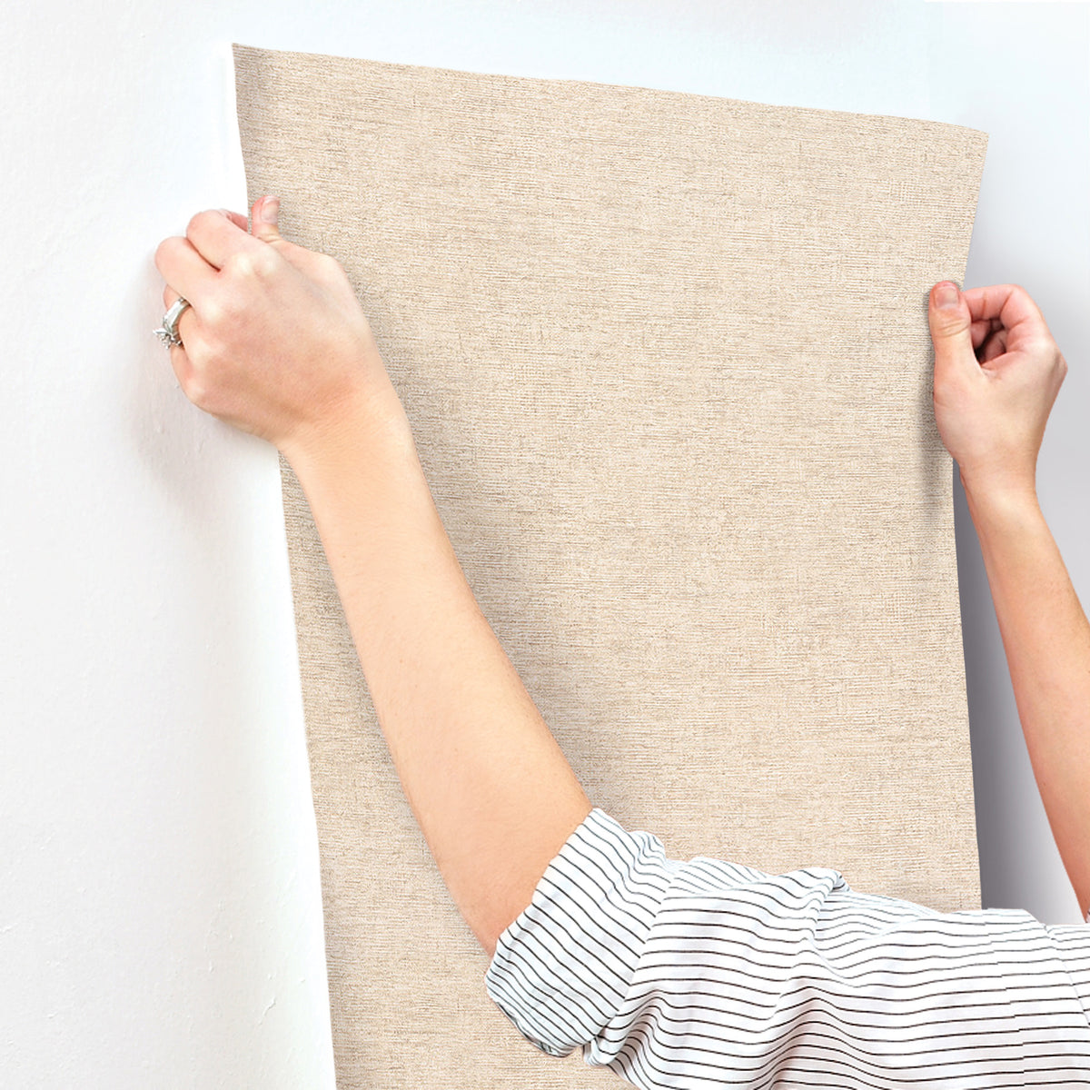 A person in a striped shirt presses a panel of Chesapeake Tansy Distressed Wallpaper by York Wallcoverings onto a white wall, smoothing it with both hands.