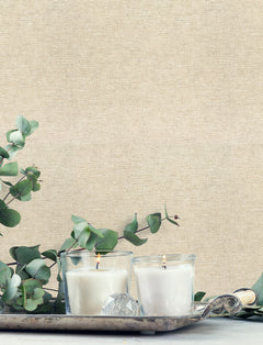 Two lit white candles in glass holders rest on a silver tray with green eucalyptus leaves, set against a neutral beige Chesapeake Tansy Distressed Wallpaper by York Wallcoverings.