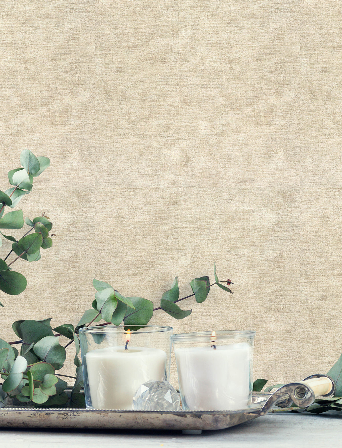Two white candles in glass holders sit on a silver tray with eucalyptus leaves, set against Chesapeake Tansy Distressed Wallpaper by York Wallcoverings. The scene is calm and minimalistic.