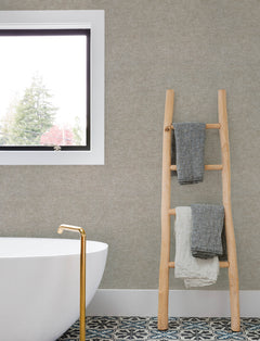 Minimalist bathroom featuring a freestanding white tub, gold faucet, patterned floor tiles, and a wooden ladder displaying two textured towels. The wall above the tub is adorned with Chesapeake Tansy Distressed Wallpaper by York Wallcoverings, with a window offering a tree view.