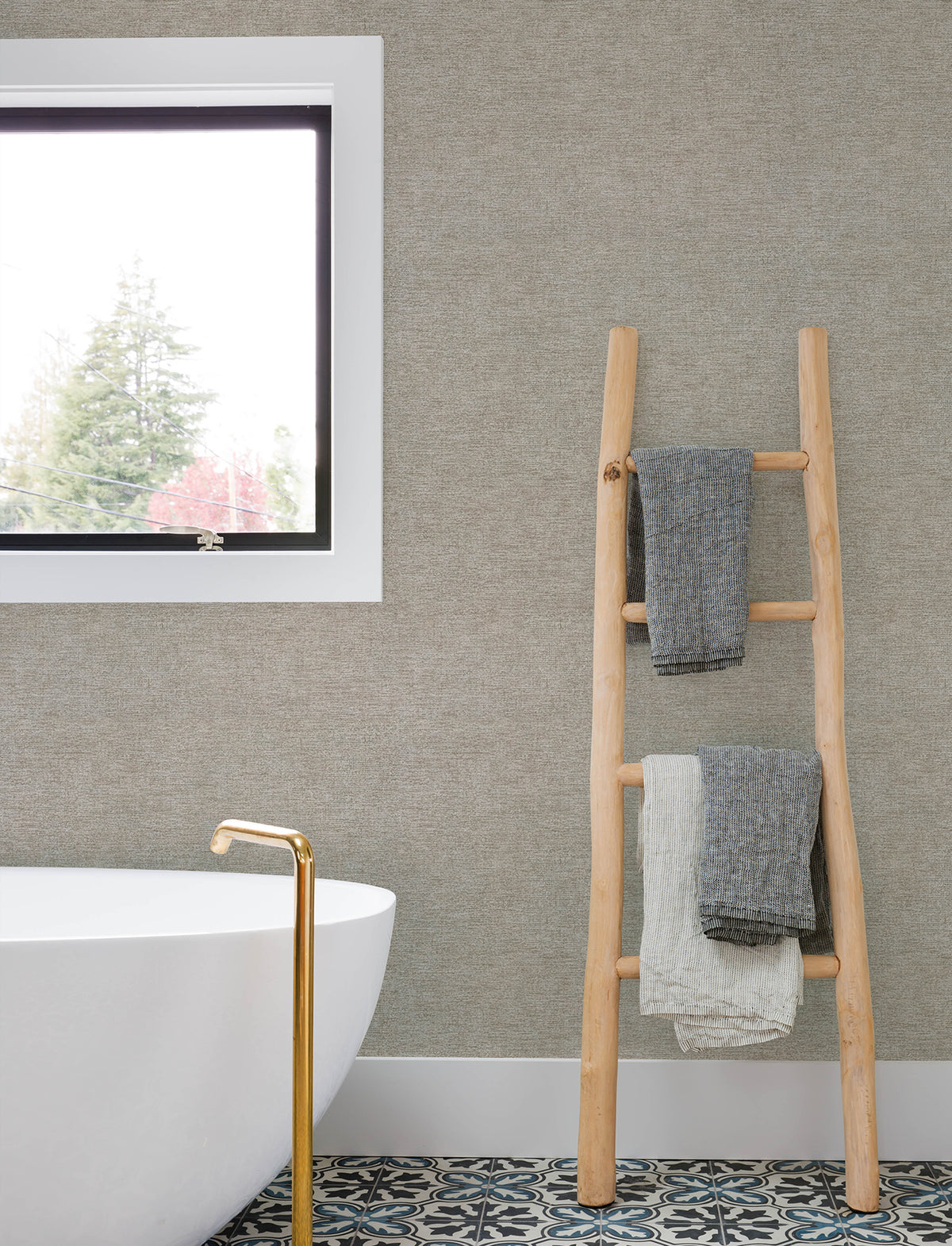 A modern bathroom featuring Chesapeake Tansy Distressed Wallpaper by York Wallcoverings, a white freestanding tub with a gold faucet, a wooden ladder holding gray and white towels, a patterned tile floor, and a window revealing trees outside.