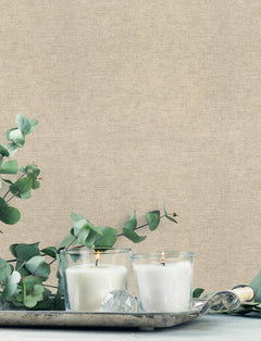 Two white candles in glass holders rest on a silver tray, surrounded by eucalyptus branches, set against York Wallcoverings’ Chesapeake Tansy Distressed Wallpaper.