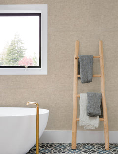 Modern bathroom with a freestanding white tub, gold faucet, patterned tile floor, wooden ladder holding three folded towels, and a window showing trees outside. The beige textured wall is adorned with Chesapeake Tansy Distressed Wallpaper by York Wallcoverings.