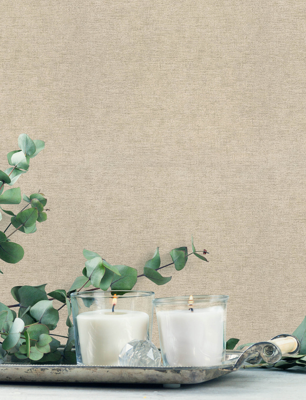 Two white candles in glass holders burn on a decorative silver tray, surrounded by eucalyptus branches, set against Chesapeake Tansy Distressed Wallpaper by York Wallcoverings.