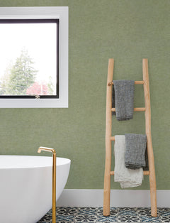 A modern bathroom with a freestanding white tub, gold faucet, patterned floor tiles, a window, and a wooden ladder holding two folded towels features York Wallcoverings’ Chesapeake Tansy Distressed Wallpaper on the green accent wall.