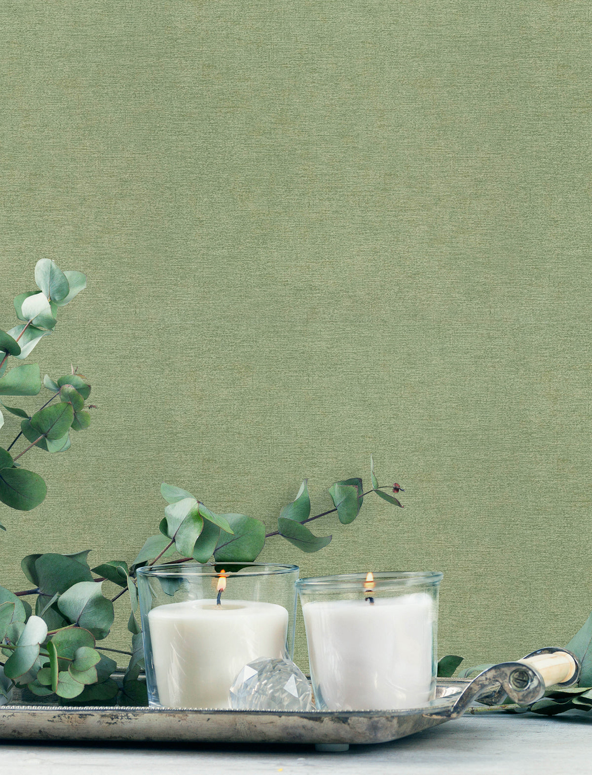 Two lit white candles in glass holders sit on a silver tray with eucalyptus branches and a decorative crystal, set against York Wallcoverings’ Chesapeake Tansy Distressed Wallpaper.