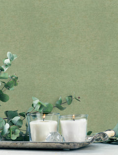 Two lit white candles in glass holders sit on a silver tray with eucalyptus branches and a decorative crystal, set against York Wallcoverings’ Chesapeake Tansy Distressed Wallpaper.