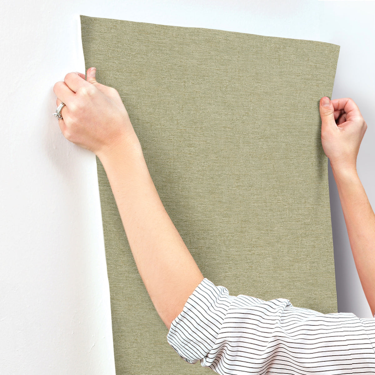 A person with a striped sleeve smooths a sheet of Chesapeake Tansy Distressed Wallpaper by York Wallcoverings onto a white wall, using both hands to align it.