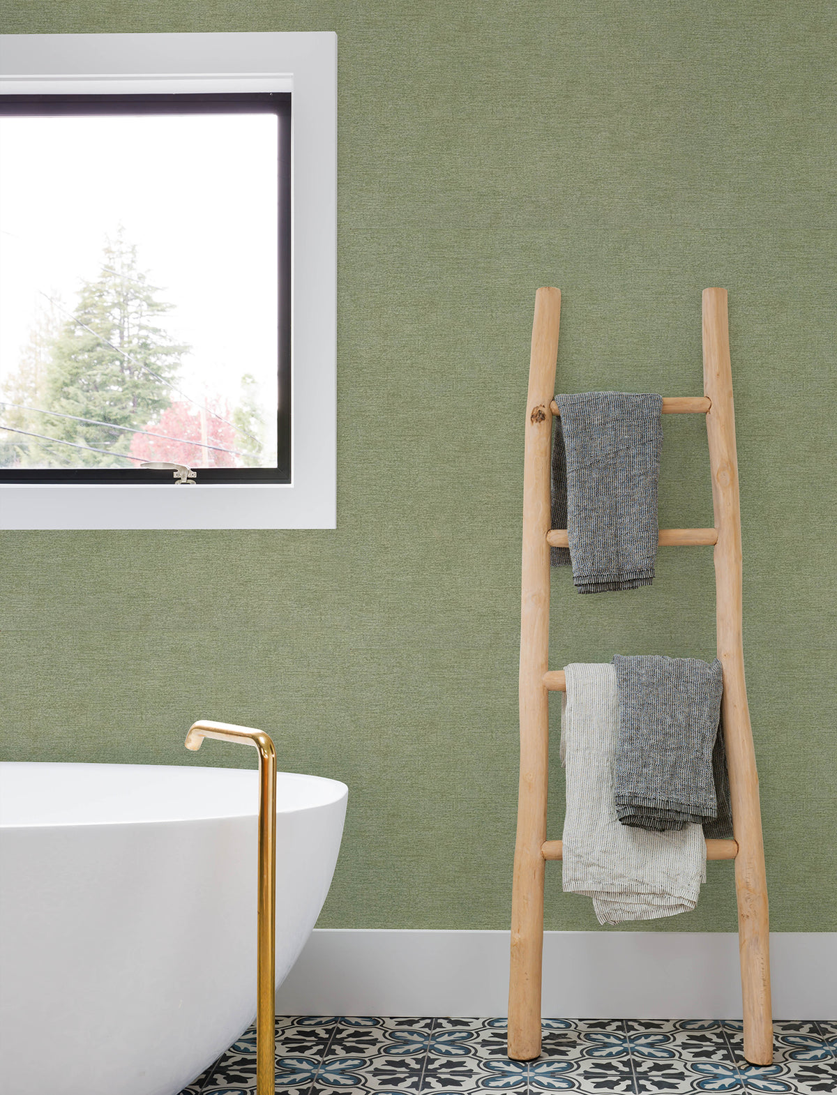 A modern bathroom featuring a freestanding white tub, gold faucet, patterned tile floor, and a window-side wooden ladder holding three folded towels, with the green wall adorned in Chesapeake Tansy Distressed Wallpaper by York Wallcoverings.