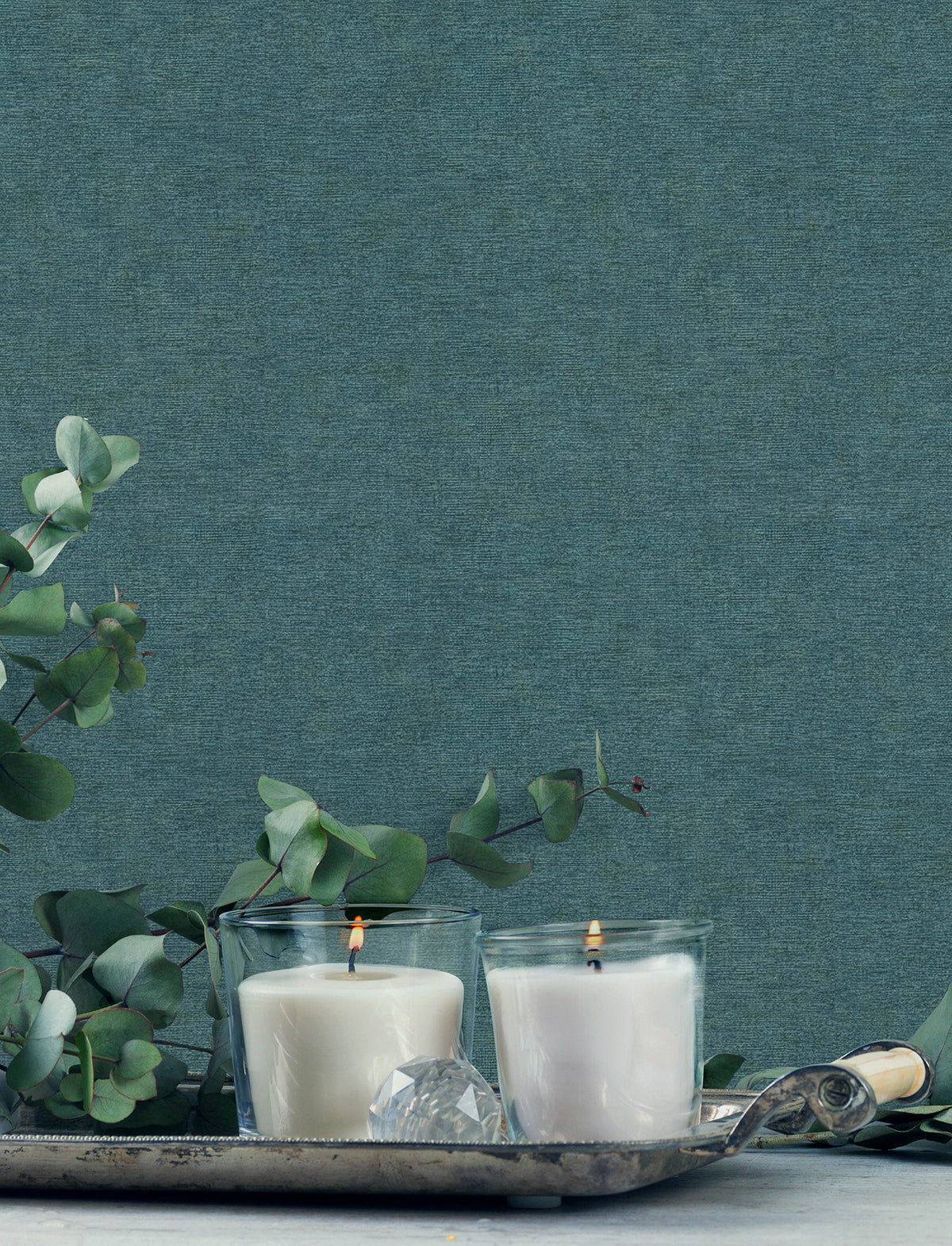 Two white candles in glass holders are lit on a silver tray, surrounded by eucalyptus branches, with York Wallcoverings Chesapeake Tansy Distressed Wallpaper as the blue textured background.