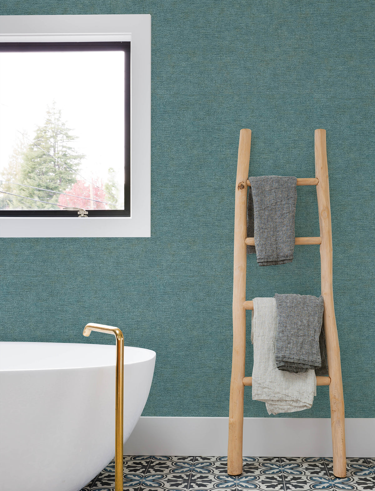 A modern bathroom featuring a white tub, gold faucet, patterned floor, wooden ladder with folded towels, a window overlooking trees, and walls adorned with Chesapeake Tansy Distressed Wallpaper by York Wallcoverings.