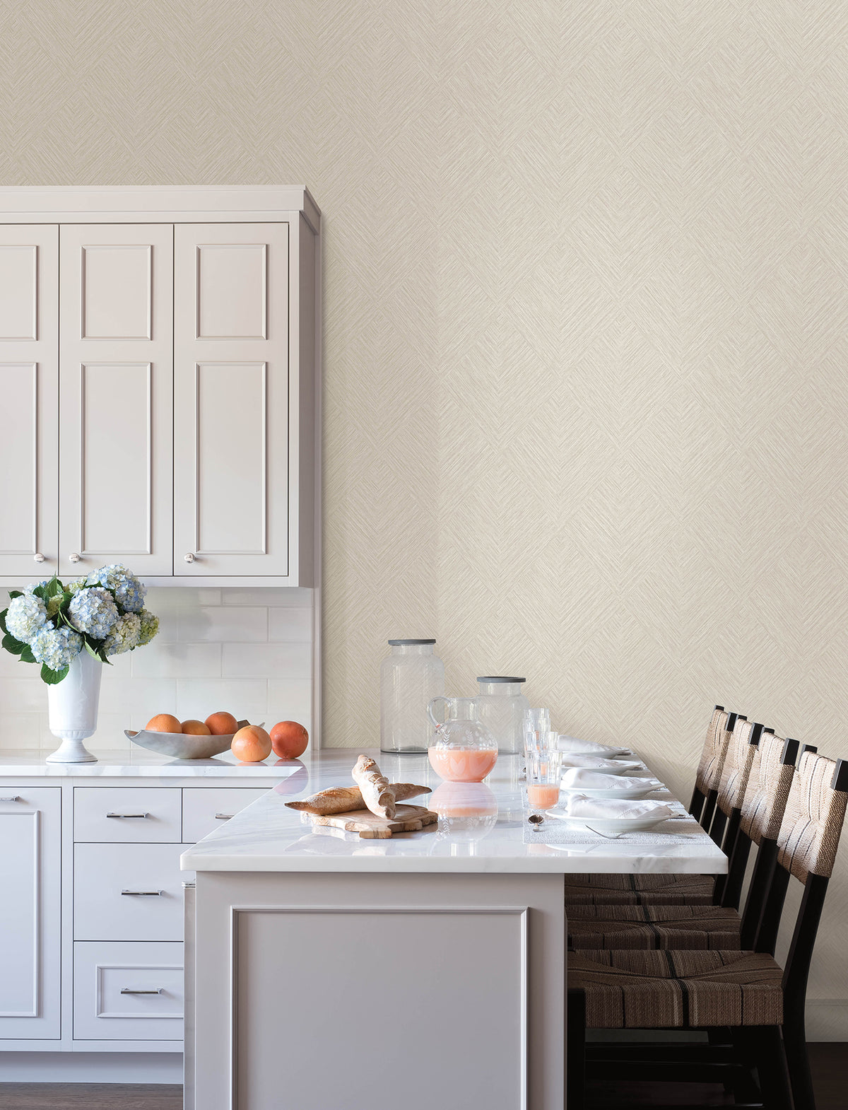 A bright kitchen featuring Chesapeake Hollis Textured Diamond Wallpaper by York Wallcoverings, with white cabinets, a marble island, four wooden chairs, a vase of flowers, jars, a glass pitcher filled with pink juice, glassware, croissants, oranges, and waffles on the counter.