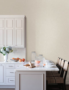 A bright kitchen featuring Chesapeake Hollis Textured Diamond Wallpaper by York Wallcoverings, with white cabinets, a marble island, four wooden chairs, a vase of flowers, jars, a glass pitcher filled with pink juice, glassware, croissants, oranges, and waffles on the counter.