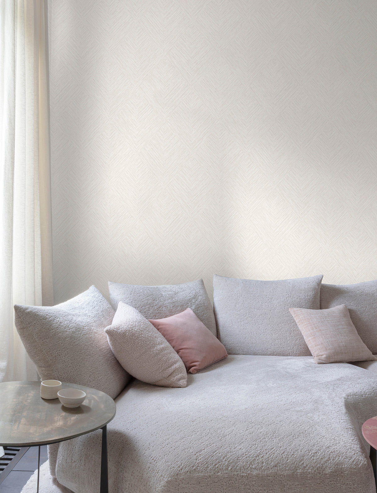 The serene space features Chesapeake Hollis Textured Diamond Wallpaper by York Wallcoverings, complementing a cozy, light-toned sofa with textured upholstery. Soft pink and gray throw pillows add warmth, while a small round side table with two white cups rests nearby. Sheer curtains filter gentle natural light, enhancing the calm and inviting ambiance.