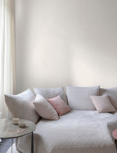 The serene space features Chesapeake Hollis Textured Diamond Wallpaper by York Wallcoverings, complementing a cozy, light-toned sofa with textured upholstery. Soft pink and gray throw pillows add warmth, while a small round side table with two white cups rests nearby. Sheer curtains filter gentle natural light, enhancing the calm and inviting ambiance.