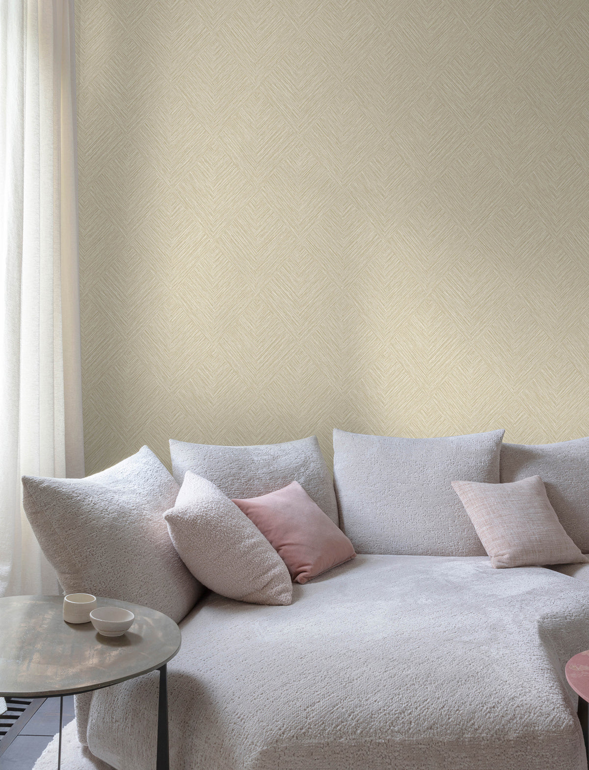 A modern living room features Chesapeake Hollis Textured Diamond Wallpaper by York Wallcoverings, adding a subtle pattern to the walls, along with a light gray textured sofa with pink and gray throw pillows, a round side table with two cups, and sheer white curtains letting in natural light.