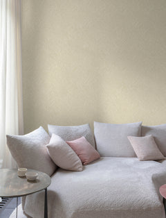 A modern living room features Chesapeake Hollis Textured Diamond Wallpaper by York Wallcoverings, adding a subtle pattern to the walls, along with a light gray textured sofa with pink and gray throw pillows, a round side table with two cups, and sheer white curtains letting in natural light.
