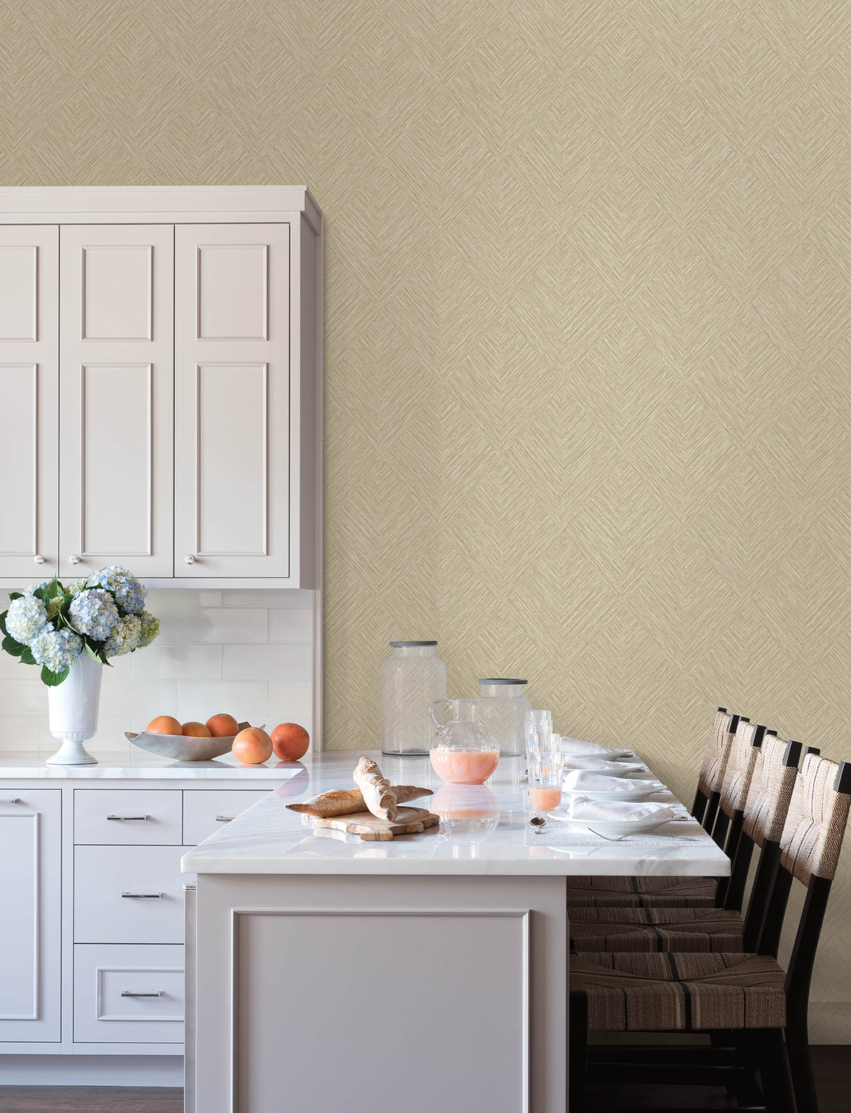 A bright kitchen features white cabinets, a marble island surrounded by four chairs, glassware, a vase of flowers, and breakfast items like juice, croissants, and fruit. The wall is adorned with Chesapeake Hollis Textured Diamond Wallpaper by York Wallcoverings in a light beige herringbone pattern.