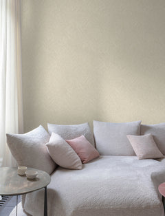 A cozy living room corner features a light gray textured sofa, pink and gray cushions, a round side table with two cups, and sheer white curtains by the window. The wall is adorned with Chesapeake Hollis Textured Diamond Wallpaper from York Wallcoverings.