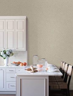 A modern kitchen features white cabinets, a marble countertop, and four barstools, with breakfast items such as juice, croissants, and fruit set out. A vase of flowers and glass jars decorate the counter. The wall showcases York Wallcoverings’ Chesapeake Hollis Textured Diamond Wallpaper for a subtle, textured look.