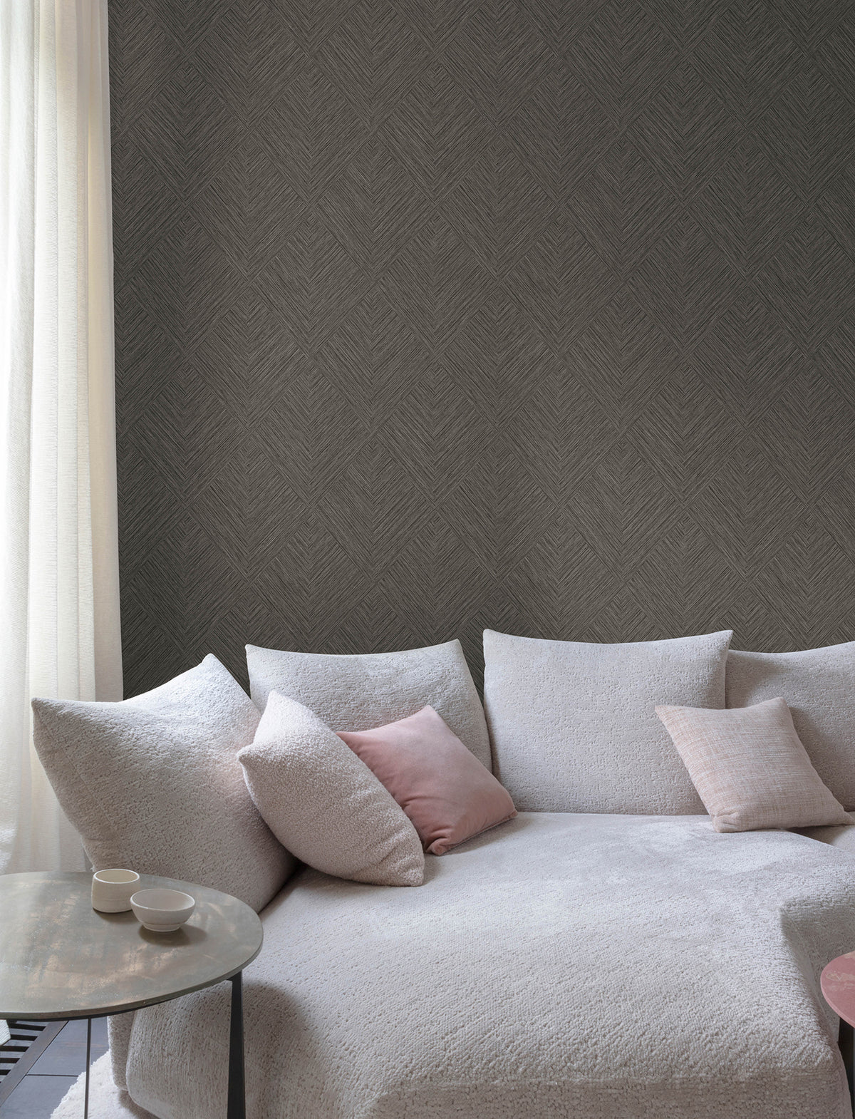 A cozy white sofa with pink and white cushions sits beside a round table holding two cups, set against a wall covered in Chesapeake Hollis Textured Diamond Wallpaper by York Wallcoverings and framed by a sheer white curtain.