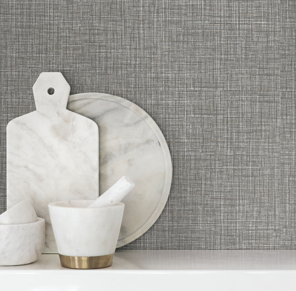 Beau Faux Fabric Wallpaper by York Wallcoverings creates a textured gray backdrop behind a white surface adorned with a marble mortar and pestle, round tray, and cutting board.