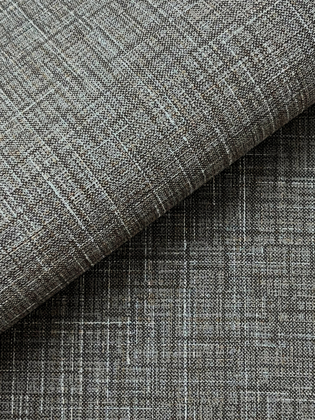 Close-up of Beau Faux Fabric Wallpaper by York Wallcoverings, showcasing a textured gray surface with a woven, crisscross pattern. One section is folded over, revealing a subtle variation in shading and texture.