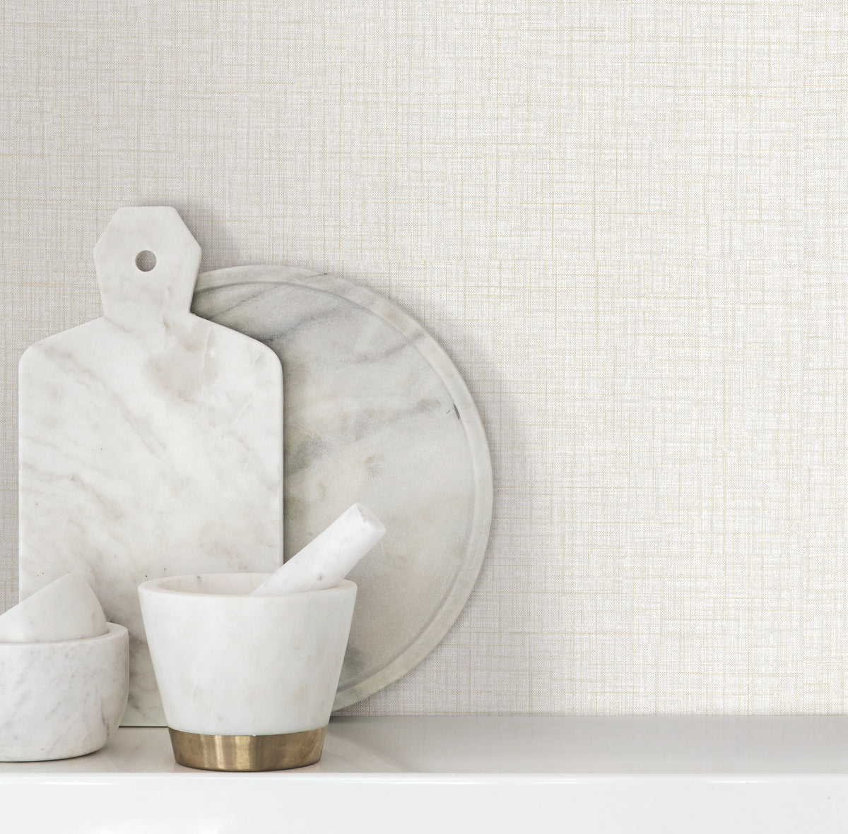 Beau Faux Fabric Wallpaper by York Wallcoverings serves as a light, textured backdrop for marble kitchenware, including a cutting board, round platter, and mortar and pestle arranged on a white surface.
